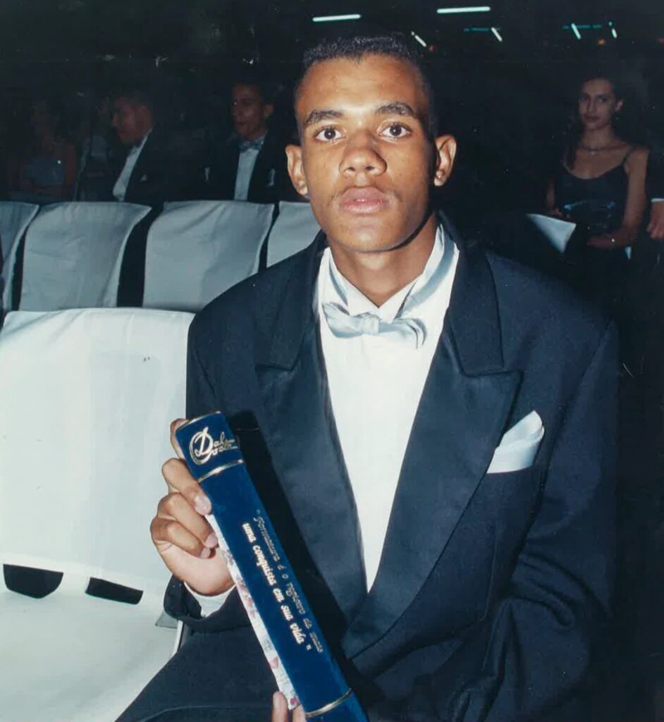 Alexandre Menezes dos Santos, jovem, de smoking preto, camisa branca e gravata-borboleta azul-clara, sentado numa cadeira coberta de tecido branco, segurando um canudo de formatura azul