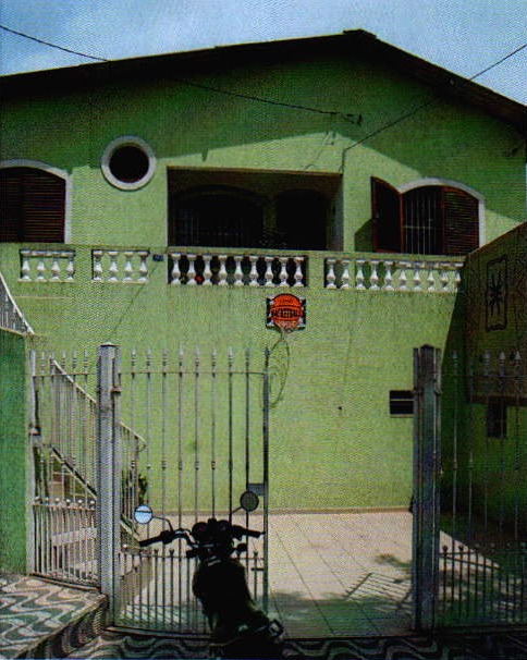 Sobrado verde com sacada de balaústres, escada externa à esquerda e portão de grade de metal na frente; atrás do portão, uma moto