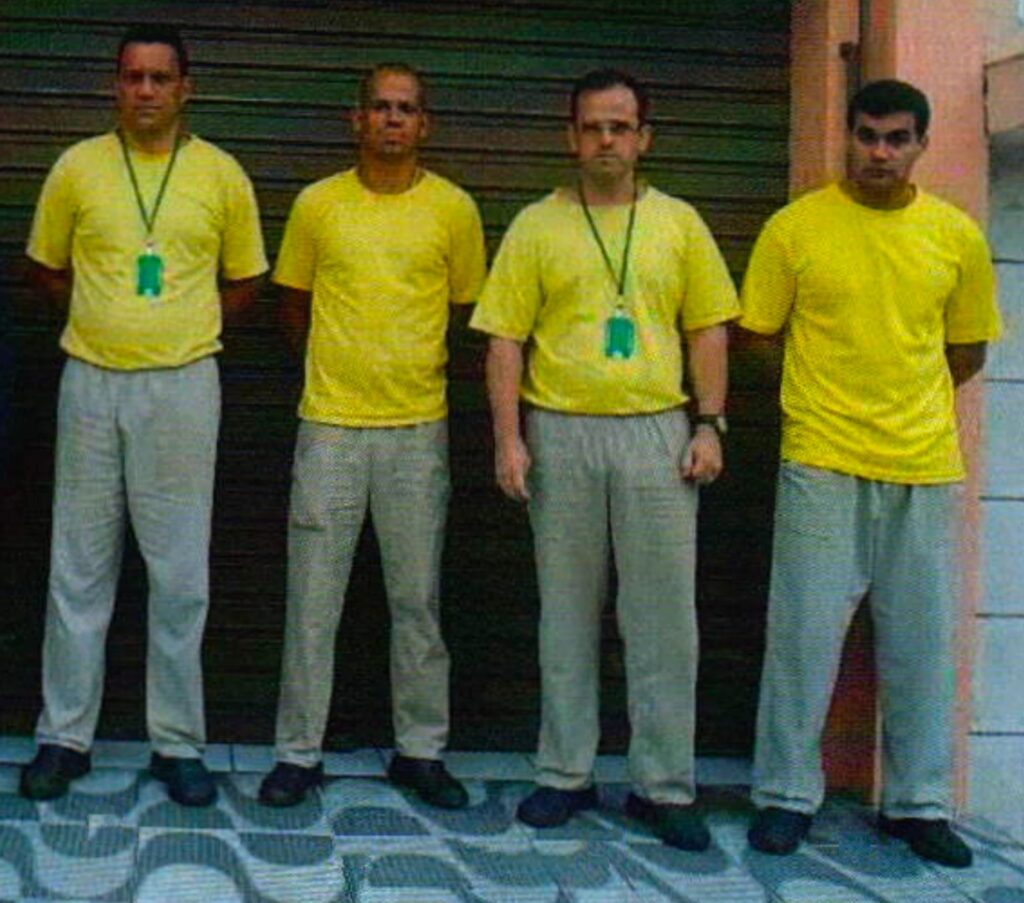 Quatro homens de camiseta amarela e calça cinza, em pé lado a lado diante de uma porta de aço: os policiais militares Carlos Magno dos Santos Diniz, Márcio Barra da Rocha, Ricardo José Manso Monteiro e Alex Sandro Soares Machado, em 2012