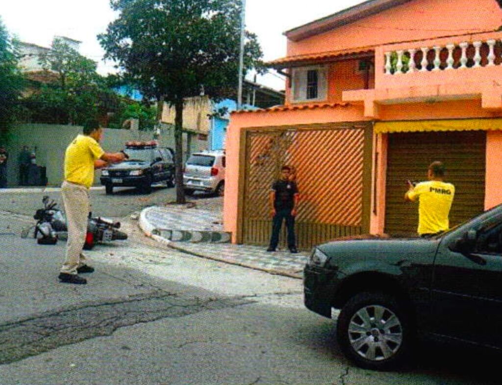 Rua com uma moto caída no asfalto. Um homem de camiseta amarela aponta as mãos como se segurasse uma arma para um homem de camisa preta parado diante de um portão de grade, enquanto outro homem de camiseta amarela faz o mesmo junto à porta de aço ao lado
