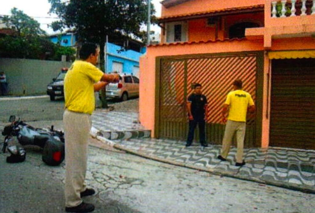 Um homem de camiseta amarela estende o braço para um homem de camisa preta diante de um portão de grade; outro homem de camiseta amarela, de costas, mira de longe; uma moto está caída na rua