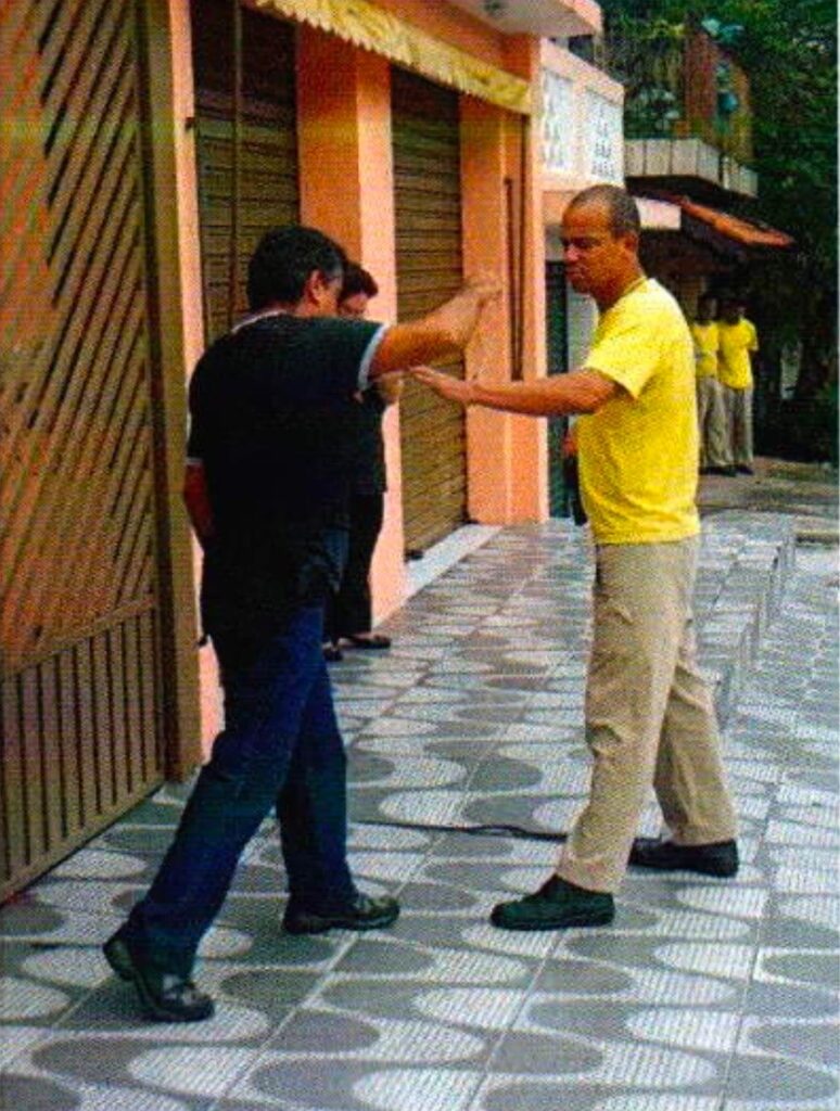 Na calçada, um homem de camisa preta estende o braço em direção ao rosto de um homem careca de camiseta amarela