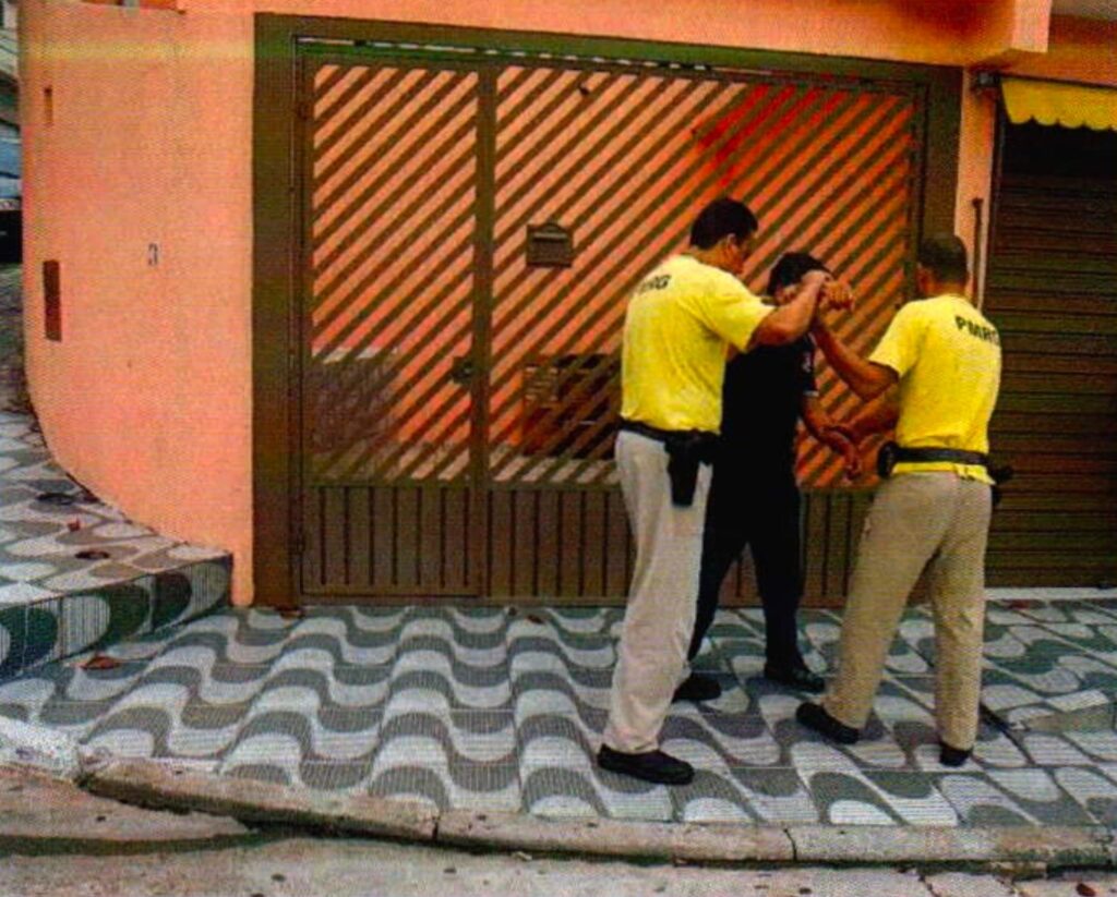 Dois homens de camiseta amarela, com coldres na cintura, seguram pelos braços um homem de camisa preta encostado em um portão de grade