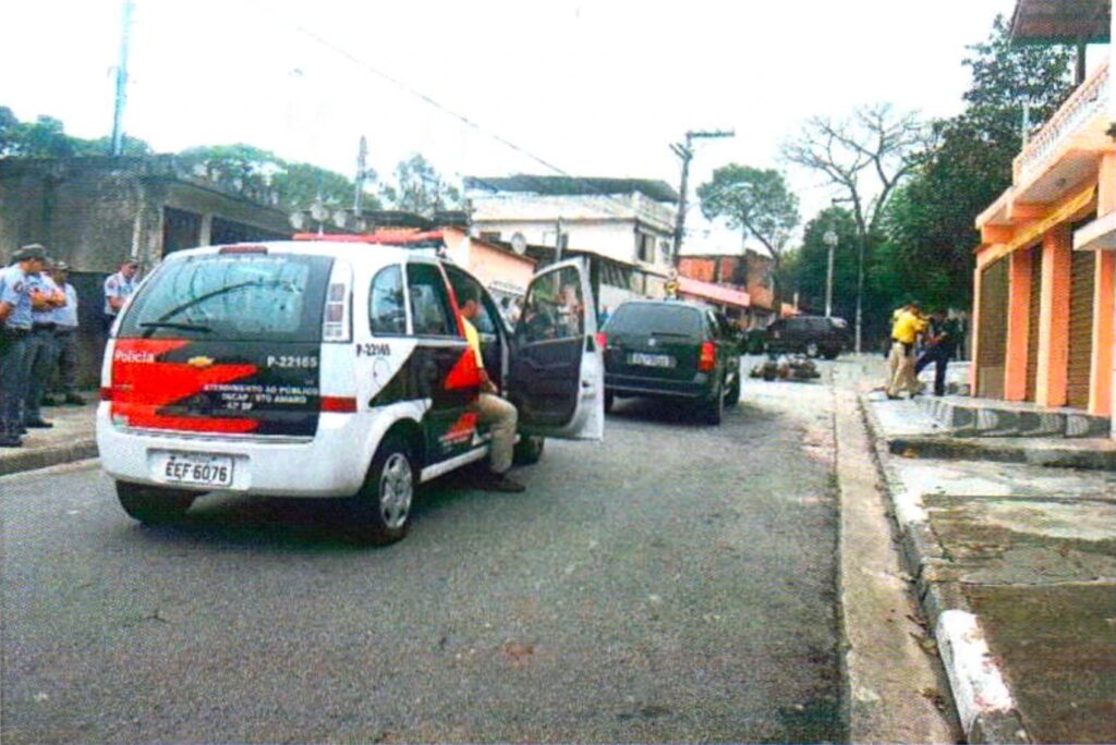 Viatura da Polícia Militar parada na rua com a porta aberta e um homem de camiseta amarela sentado no banco; à esquerda, policiais militares fardados observam; ao fundo, na calçada, a encenação diante da casa