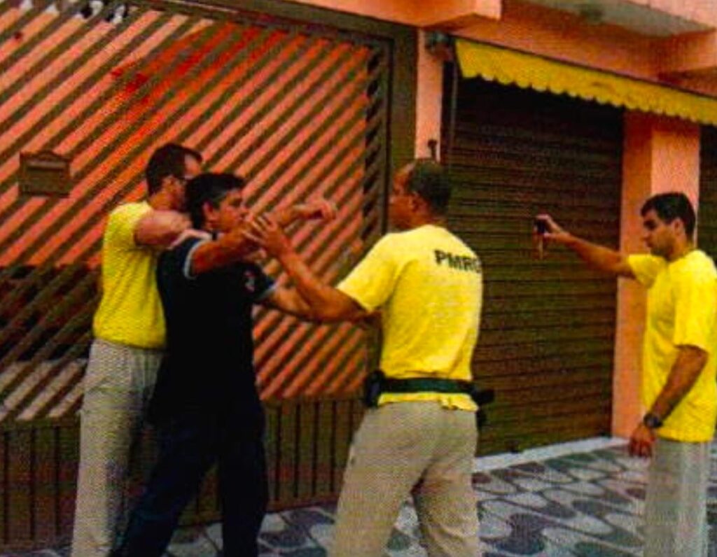 Diante de um portão de grade, um homem de camiseta amarela prende pelo pescoço, por trás, um homem de camisa preta; outro de camiseta amarela o segura pela frente e um terceiro, à direita, aponta um frasco de spray