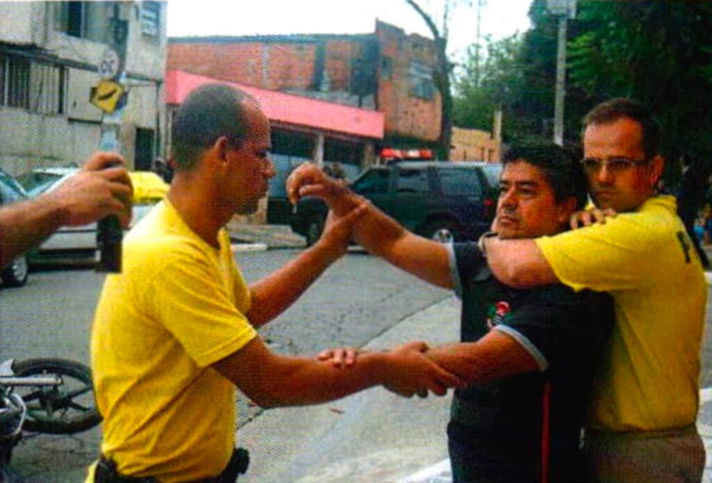 Um homem de óculos e camiseta amarela prende pelo pescoço, por trás, um homem de camisa preta; à frente, um homem careca de camiseta amarela segura o braço dele