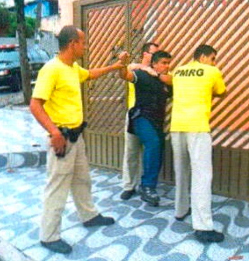 Junto a um portão de grade, três homens de camiseta amarela cercam um homem de camisa preta; o da esquerda, com coldre na cintura, segura o braço dele