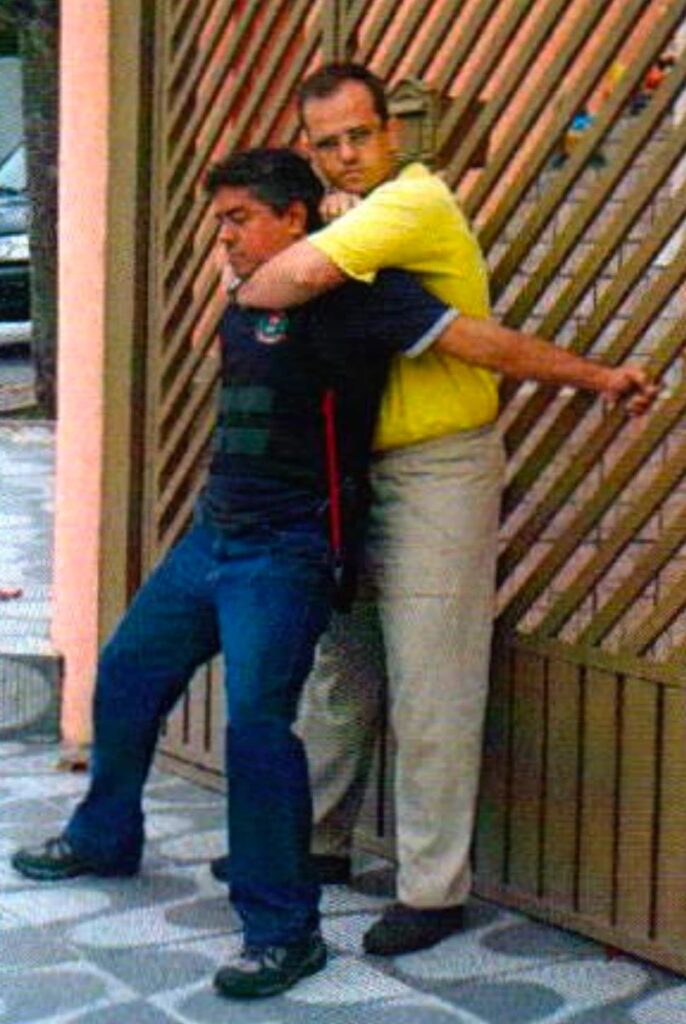 Diante de um portão de grade, um homem de óculos e camiseta amarela prende pelo pescoço, por trás, um homem de camisa preta, que estica os braços para trás e segura a grade