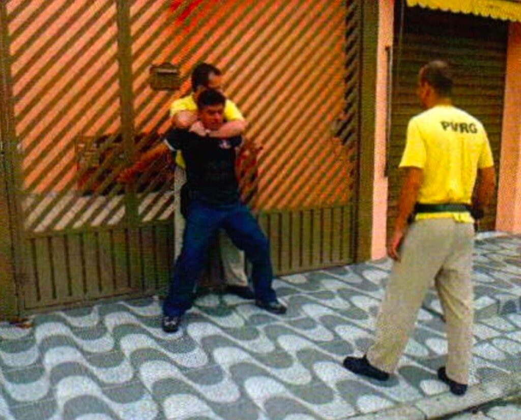 Junto a um portão de grade, um homem de camiseta amarela prende pelo pescoço um homem de camisa preta; à direita, de costas, um homem careca de camiseta amarela se afasta