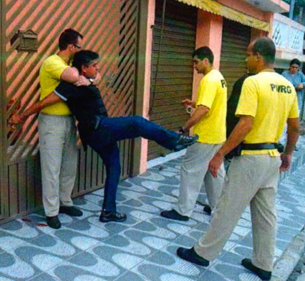Na calçada, um homem de óculos e camiseta amarela prende pelo pescoço um homem de camisa preta, que levanta a perna num chute em direção a outro homem de camiseta amarela; à direita, um terceiro, de costas