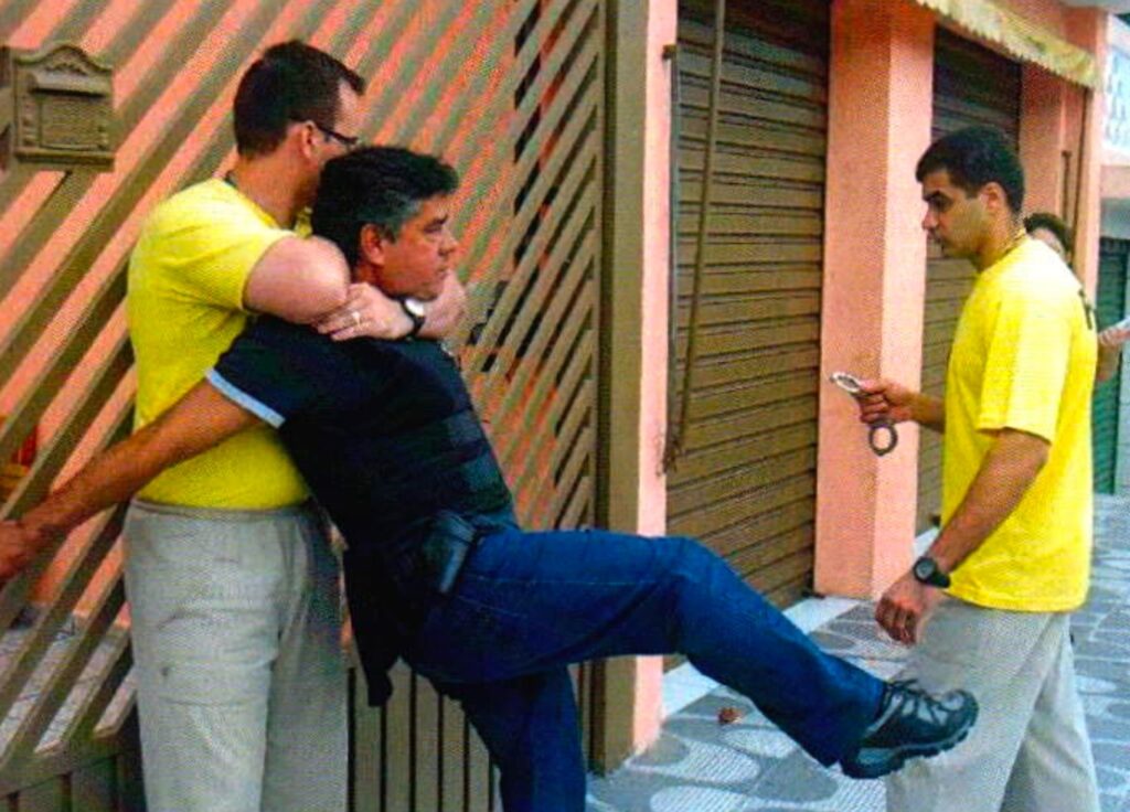 Um homem de óculos e camiseta amarela prende pelo pescoço um homem de camisa preta, que estica a perna num chute; à direita, um homem jovem de camiseta amarela segura algemas