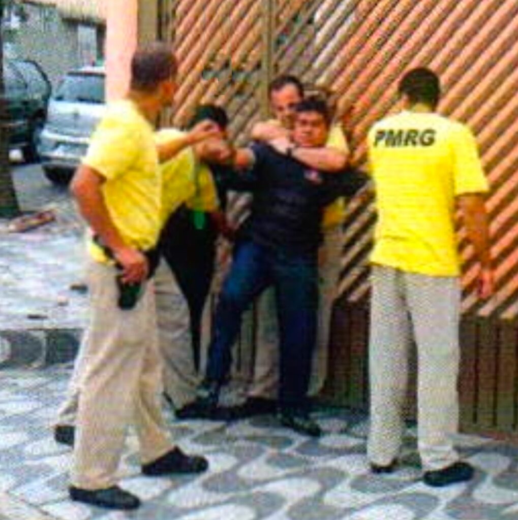 Junto a um portão de grade, um homem de camisa preta preso pelo pescoço por trás estende o braço em direção a um homem careca de camiseta amarela; outros dois homens de camiseta amarela estão ao lado