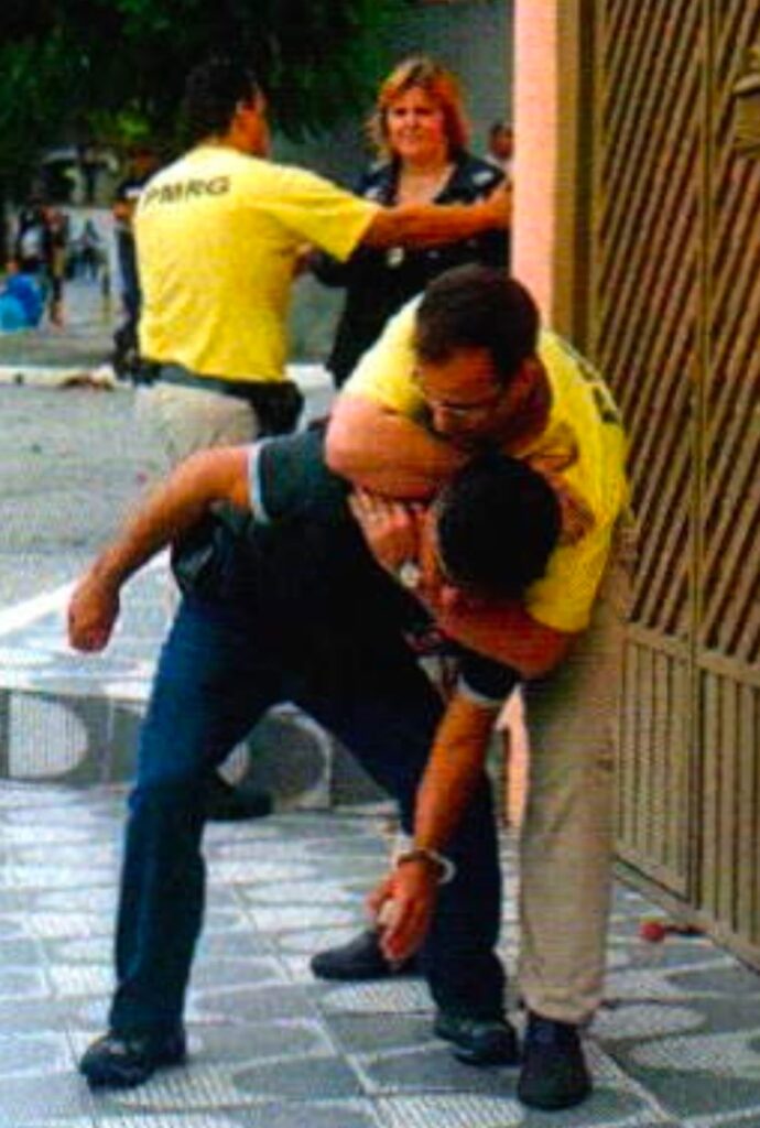 Um homem de óculos e camiseta amarela prende pelo pescoço e curva para a frente um homem de camisa preta com uma algema no pulso; ao fundo, outro homem de camiseta amarela segura uma mulher loira