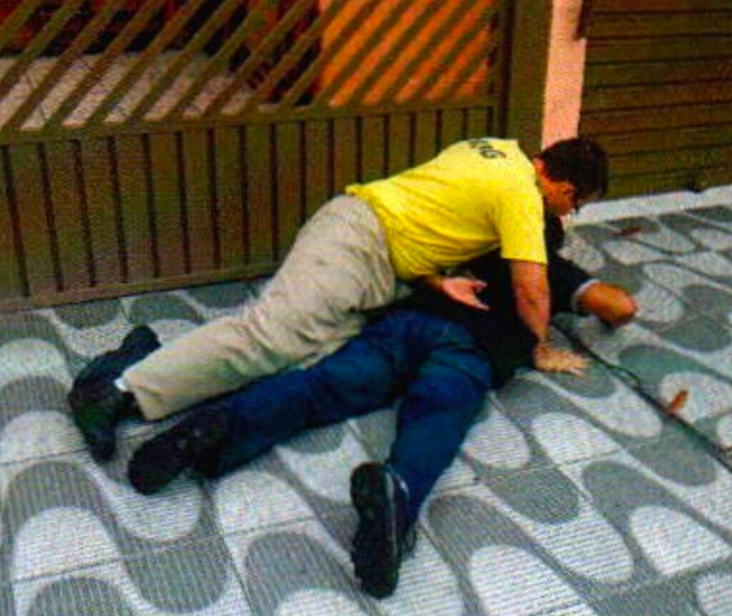 Na calçada junto a um portão, um homem de camisa preta está de bruços e um homem de camiseta amarela está deitado por cima dele