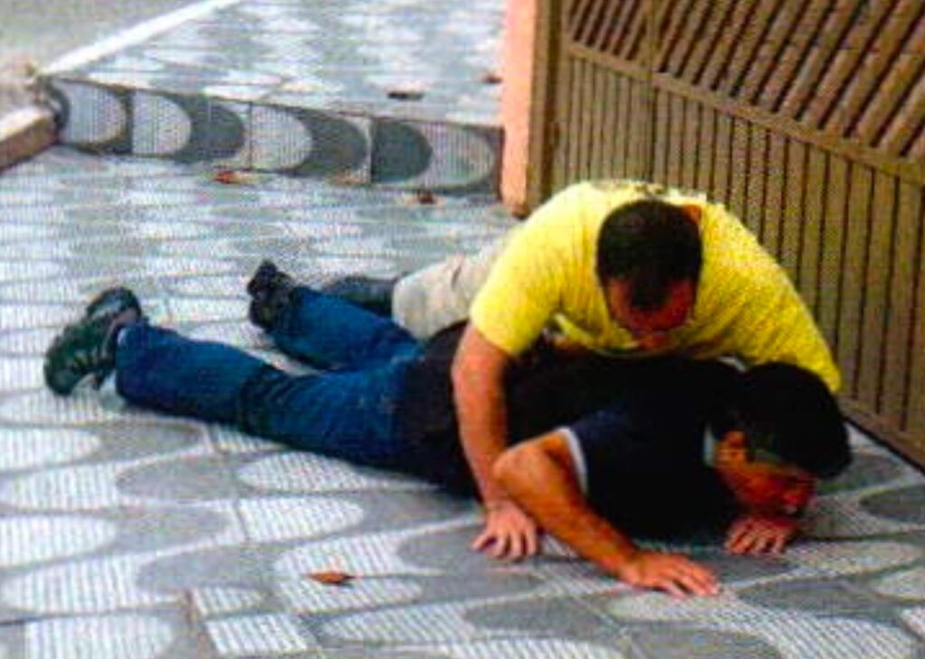 Um homem de camiseta amarela está por cima de um homem de camisa preta e calça jeans, deitado de bruços na calçada diante de um portão de grade