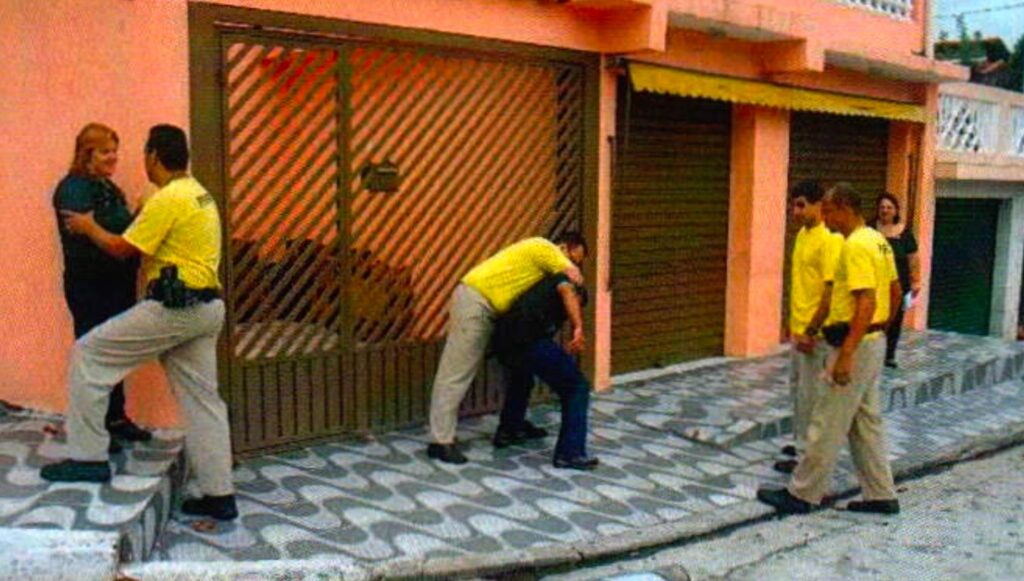 Fachada com portão de grade: à esquerda, um homem de camiseta amarela segura uma mulher loira; no centro, outro prende pelo pescoço um homem de camisa preta curvado; à direita, dois homens de camiseta amarela observam