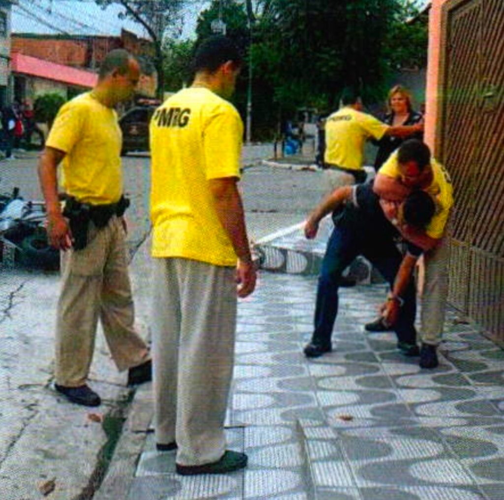 Na calçada, um homem de óculos e camiseta amarela prende pelo pescoço um homem de camisa preta curvado para a frente; dois homens de camiseta amarela observam em primeiro plano; ao fundo, outro segura uma mulher loira