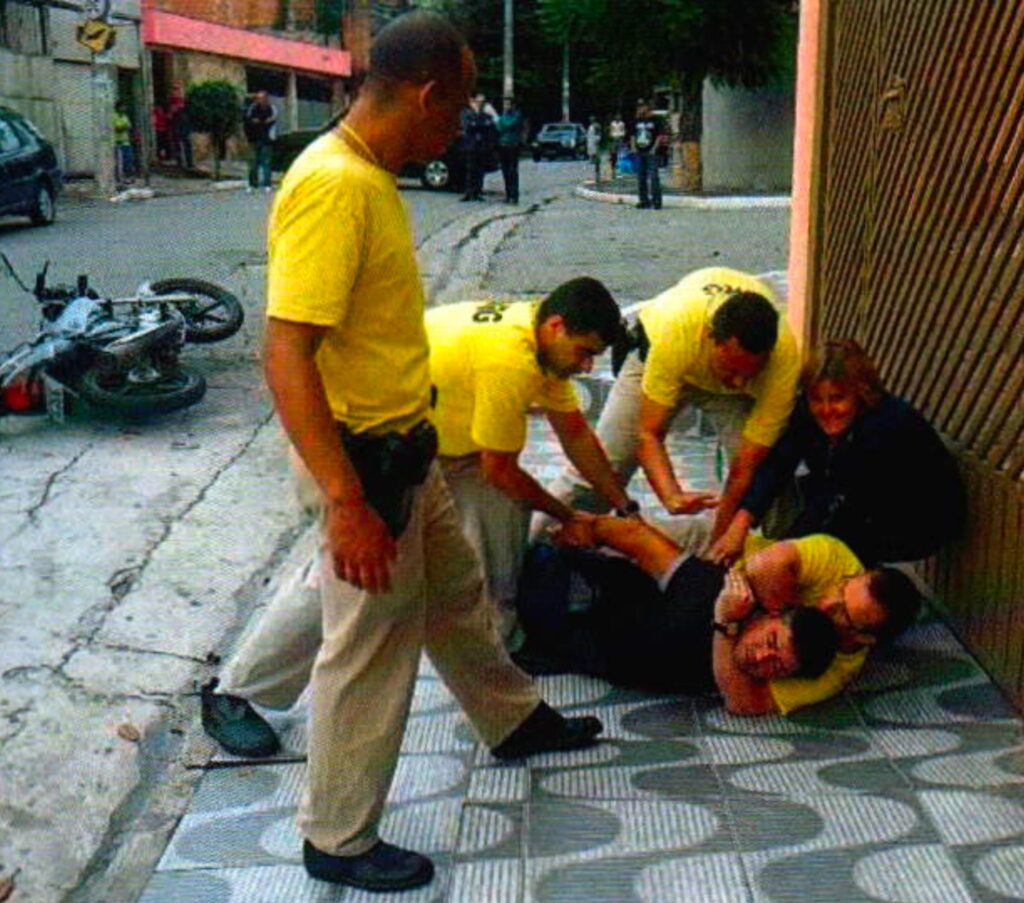 Na calçada, um homem de camisa preta está deitado, preso pelo pescoço por um homem de camiseta amarela; dois outros se inclinam sobre ele, uma mulher loira agachada o puxa e um homem careca de pé observa; ao fundo, uma moto caída