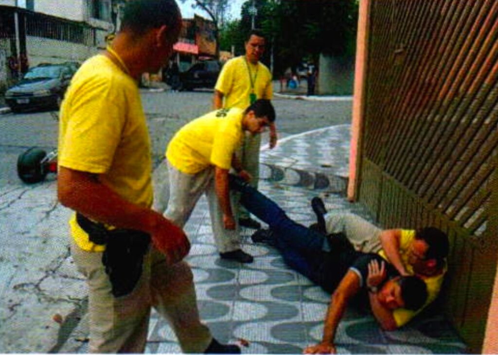 Na calçada junto a um portão, um homem de camiseta amarela deitado prende pelo pescoço um homem de camisa preta; outro se inclina sobre eles e dois observam de pé; ao fundo, uma moto caída na rua
